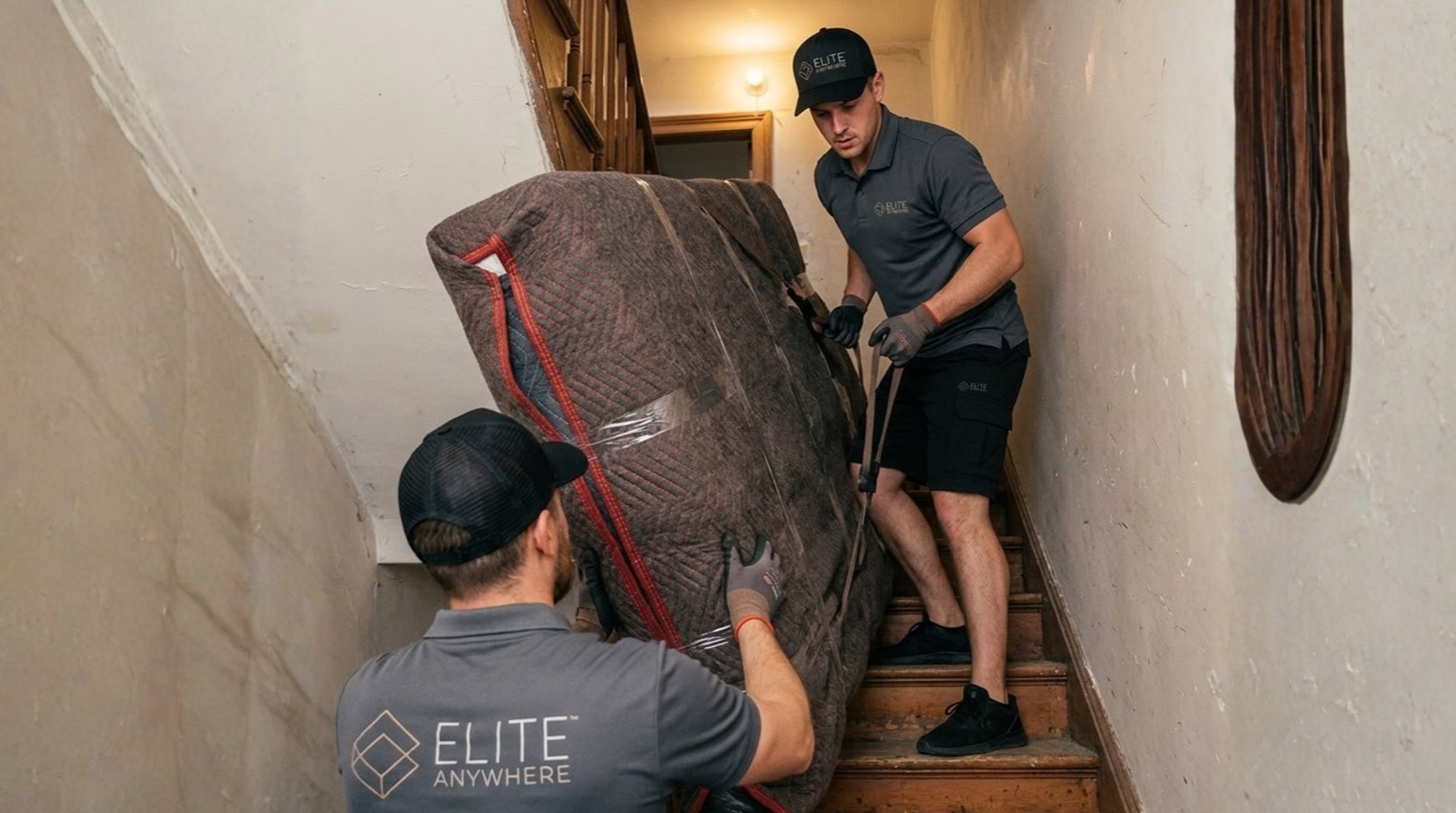 Two Elite Anywhere movers carrying a wrapped sofa down a narrow stairwell
