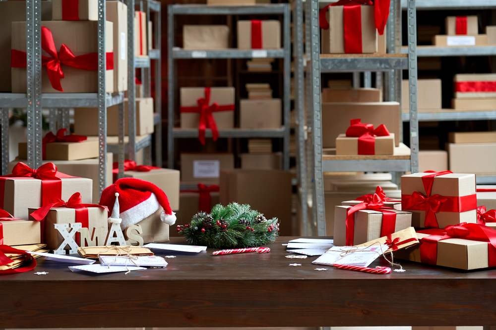 Decorated Merry Christmas table with gifts boxes in warehouse interior background. Many presents wrapped with red ribbons and letters on desk in storage. Xmas postal shipping delivery concept.